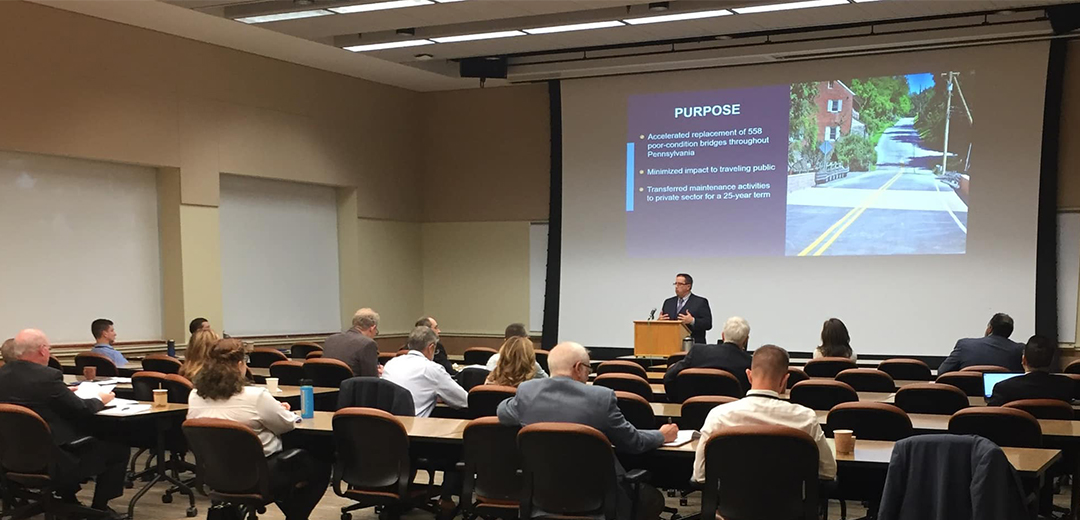 man standing at the front of a conference room with a powerpoint presentation in the background as he speaks to a crowd